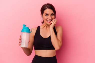Pembe arka planda izole edilmiş protein içeceği içen genç beyaz kadın tırnaklarını yiyor, gergin ve çok endişeli..