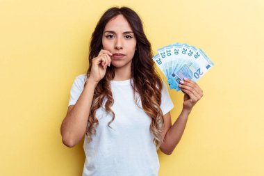 Sarı arka planda izole edilmiş banknotlar tutan genç melez bir kadın dudaklarında parmaklarıyla bir sır saklıyor..