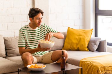 Genç melez adam kanepede oturmuş patlamış mısır yiyor.