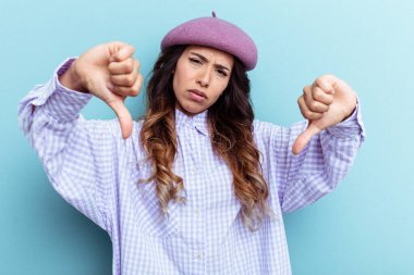 Mavi arka planda izole edilmiş genç Meksikalı kadın baş parmağını aşağı gösteriyor ve hoşlanmadığını ifade ediyor..