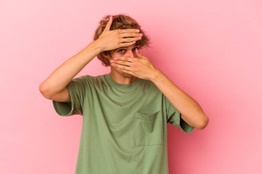 Makyajlı genç beyaz adam pembe arka planda izole edilmiş parmaklarıyla kameraya göz kırpıyor, yüzünü kapatmaktan utanıyor..