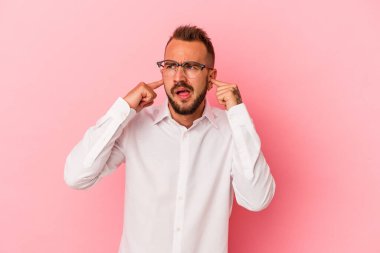 Pembe arka planda dövmeleri olan, kulaklarını parmaklarıyla kapatan, stresli ve çaresiz genç beyaz bir adam..