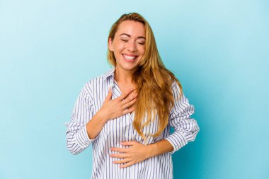 Mavi arka planda izole edilmiş beyaz kadın mutlu bir şekilde gülüyor ve ellerini karnının üstünde tutarak eğleniyor..