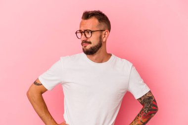Pembe arka planda izole edilmiş dövmeleri olan genç beyaz adam kafası karışmış, şüpheli ve güvensiz hissediyor..