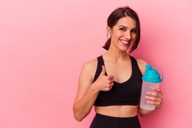 Beyaz bir kadın pembe arka planda izole edilmiş protein içeceği içiyor gülümsüyor ve başparmağını kaldırıyor.