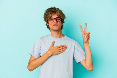 Makyajlı genç beyaz adam mavi arka planda izole edilmiş yemin ediyor, elini göğsüne koyuyor..