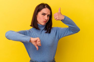 Sarı arka planda izole edilmiş genç beyaz kadın baş parmağını kaldırıyor ve baş parmak aşağı, zor seçim konsepti.