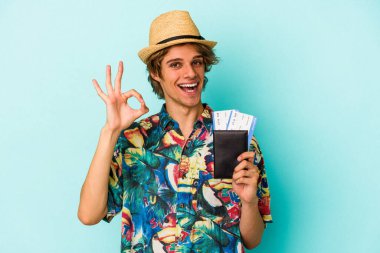 Makyajlı, pasaportlu genç beyaz adam mavi arka planda izole edilmiş neşeli ve kendinden emin bir şekilde iyi niyet gösteriyor..