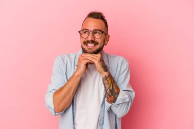 Pembe arka planda dövmeleri olan genç beyaz adam ellerini çenesinin altında tutuyor ve mutlu bir şekilde yan tarafa bakıyor..