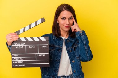 Sarı arka planda izole edilmiş bir alçıpan tutan genç beyaz kadın şakağı parmağıyla işaret ediyor, düşünüyor ve bir göreve odaklanıyor..