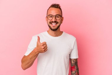 Pembe arka planda izole edilmiş, gülümseyen ve baş parmağını kaldıran beyaz dövmeli genç bir adam.