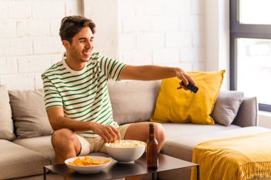 Genç melez adam kanepede oturmuş patlamış mısır yiyor.