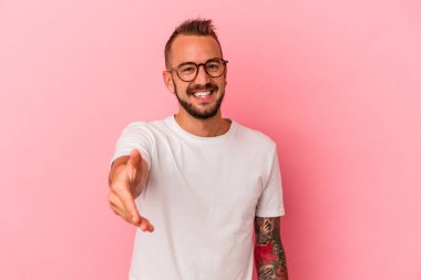 Pembe arka planda izole edilmiş beyaz dövmeli genç bir adam. Kameraya elini uzatıp selam veriyor..