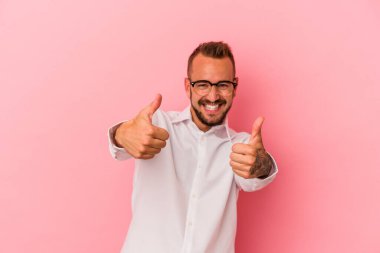 Pembe arka planda izole edilmiş beyaz dövmeli genç bir adam iki parmağını da kaldırıyor, gülümsüyor ve kendinden emin..