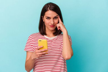 Elinde cep telefonu olan beyaz bir kadın mavi arka planda izole edilmiş. Şakağı işaret ediyor, düşünüyor ve bir göreve odaklanıyor..