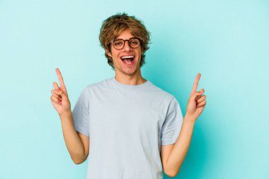 Mavi arka planda soyutlanmış beyaz makyajlı genç bir adam iki ön parmağı da boş bir alan gösteriyor..