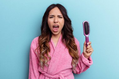 Bornoz giymiş genç Meksikalı bir kadın mavi arka planda izole edilmiş bir fırça tutuyor. Çok sinirli ve agresif bağırıyor..