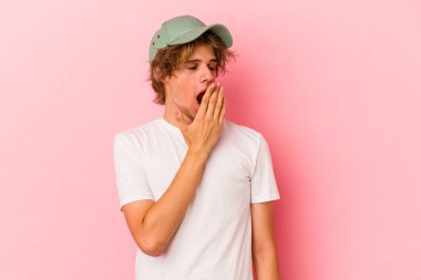 Pembe arka planda soyutlanmış beyaz makyajlı genç bir adam esneyerek ağzını eliyle kapatan yorgun bir jest yapıyor..