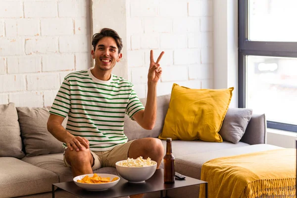 stock image Young mixed race man eating popcorns sitting on the sofa joyful and carefree showing a peace symbol with fingers.