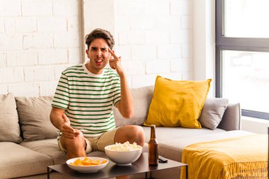 Genç melez adam kanepede oturmuş patlamış mısır yiyor işaret parmağıyla hayal kırıklığı jesti yapıyor..