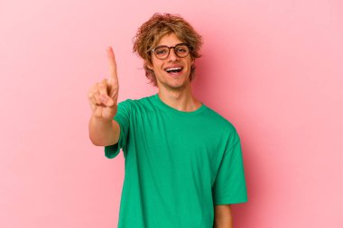 Pembe arka planda soyutlanmış beyaz makyajlı genç bir adam parmağıyla bir numarayı gösteriyor..