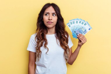 Sarı arka planda izole edilmiş banknotları tutan genç melez kadın kafası karışmış, şüpheli ve güvensiz hissediyor..