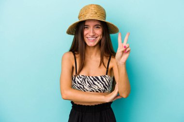 Genç beyaz kadın kumsala gidiyor. Mavi arka planda izole edilmiş bikini giyiyor. İki numarayı parmaklarıyla gösteriyor..