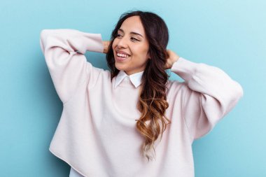 Genç Meksikalı kadın mavi arka planda izole edilmiş. Kendine güveniyor, elleri başının arkasında..