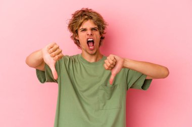 Pembe arka planda soyutlanmış beyaz tenli genç bir adam baş parmağını aşağı gösteriyor ve hoşlanmadığını ifade ediyor..