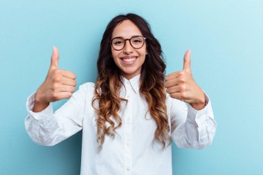 Mavi arka planda izole edilmiş genç Meksikalı kadın gülümsüyor ve başparmağını kaldırıyor.