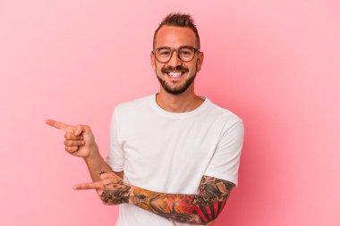 Pembe arka planda izole edilmiş beyaz dövmeli genç bir adam işaret parmağıyla işaret ederek heyecanlanıyor..