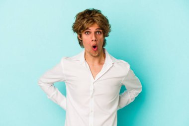 Makyajlı genç beyaz adam mavi arka planda izole edilmiş. Gördüğü bir şey yüzünden şoka uğruyor..