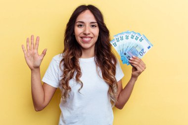 Sarı arka planda izole edilmiş banknotları tutan genç melez kadın gülümseyerek beş numarayı parmaklarıyla gösteriyor..