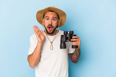 Dürbünü mavi arka planda tutan beyaz dövmeli genç adam şaşırdı ve şok oldu..