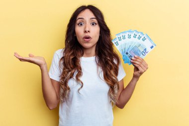 Sarı arka planda izole edilmiş banknotlar tutan genç melez kadın omuz silker ve gözleri açık kalır..