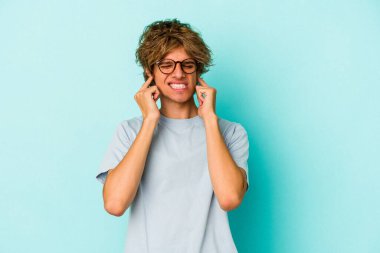 Makyajlı genç beyaz adam mavi arka planda izole edilmiş kulakları elleriyle kapatıyor..