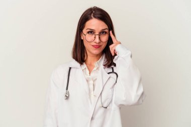 Beyaz arka planda izole edilmiş genç bir doktor şakağı parmağıyla işaret ediyor, düşünüyor ve bir göreve odaklanıyor..