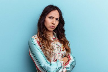 Mavi arka planda izole edilmiş genç Meksikalı kadın hoşnutsuzca kaşlarını çatmış, kollarını kavuşturmuş..