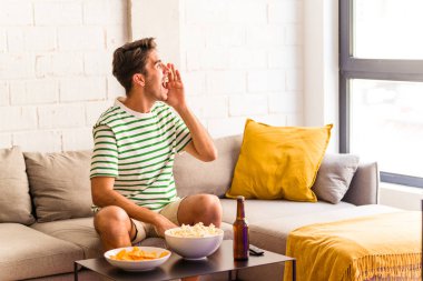 Genç melez adam kanepede oturmuş patlamış mısır yiyor bağırıyor ve avuç içini açıyor..