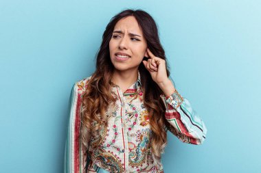 Mavi arka planda izole edilmiş genç Meksikalı kadın kulaklarını elleriyle kapatıyor..