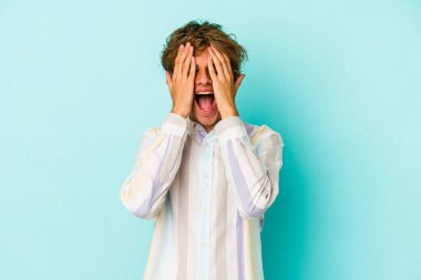 Mavi arka planda soyutlanmış beyaz tenli genç adam yüzünün yarısını avuç içiyle kaplayarak eğleniyor..