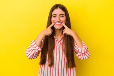 Sarı arka planda izole edilmiş genç beyaz kadın gülümsüyor, parmaklarını ağzına doğrultuyor..