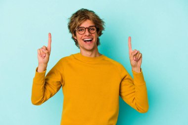 Mavi arka planda soyutlanmış beyaz makyajlı genç bir adam iki ön parmağı da boş bir alan gösteriyor..