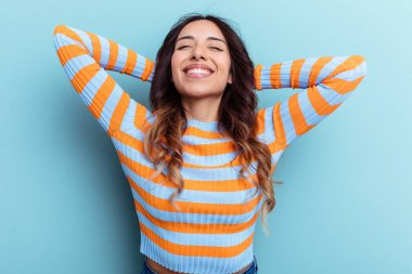 Genç Meksikalı kadın mavi arka planda izole edilmiş. Kendine güveniyor, elleri başının arkasında..