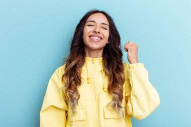 Mavi arka planda izole edilmiş genç Meksikalı kadın zaferi, tutkuyu ve coşkuyu kutluyor, mutlu bir ifade..