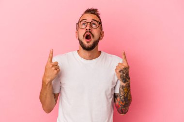 Pembe arka planda izole edilmiş, ağzı açık, üst tarafı işaret eden dövmeleri olan beyaz bir genç..