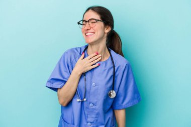 Mavi arka planda izole edilmiş genç beyaz hemşire elini göğsünde tutarak kahkahalar atıyor..