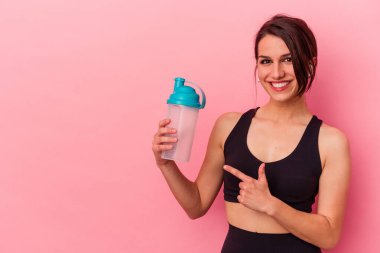 Beyaz bir kadın pembe arka planda izole edilmiş protein içeceği içiyor gülümsüyor ve boş alanda bir şeyler gösteriyor..