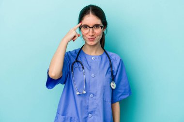 Kafkasyalı genç hemşire mavi arka planda izole edildi. Şakağı parmağıyla göstererek, düşünerek, bir göreve odaklandı..