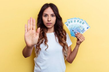 Sarı arka planda izole edilmiş banknotları tutan genç melez kadın elini uzatıp dur işareti göstererek seni engelliyor..